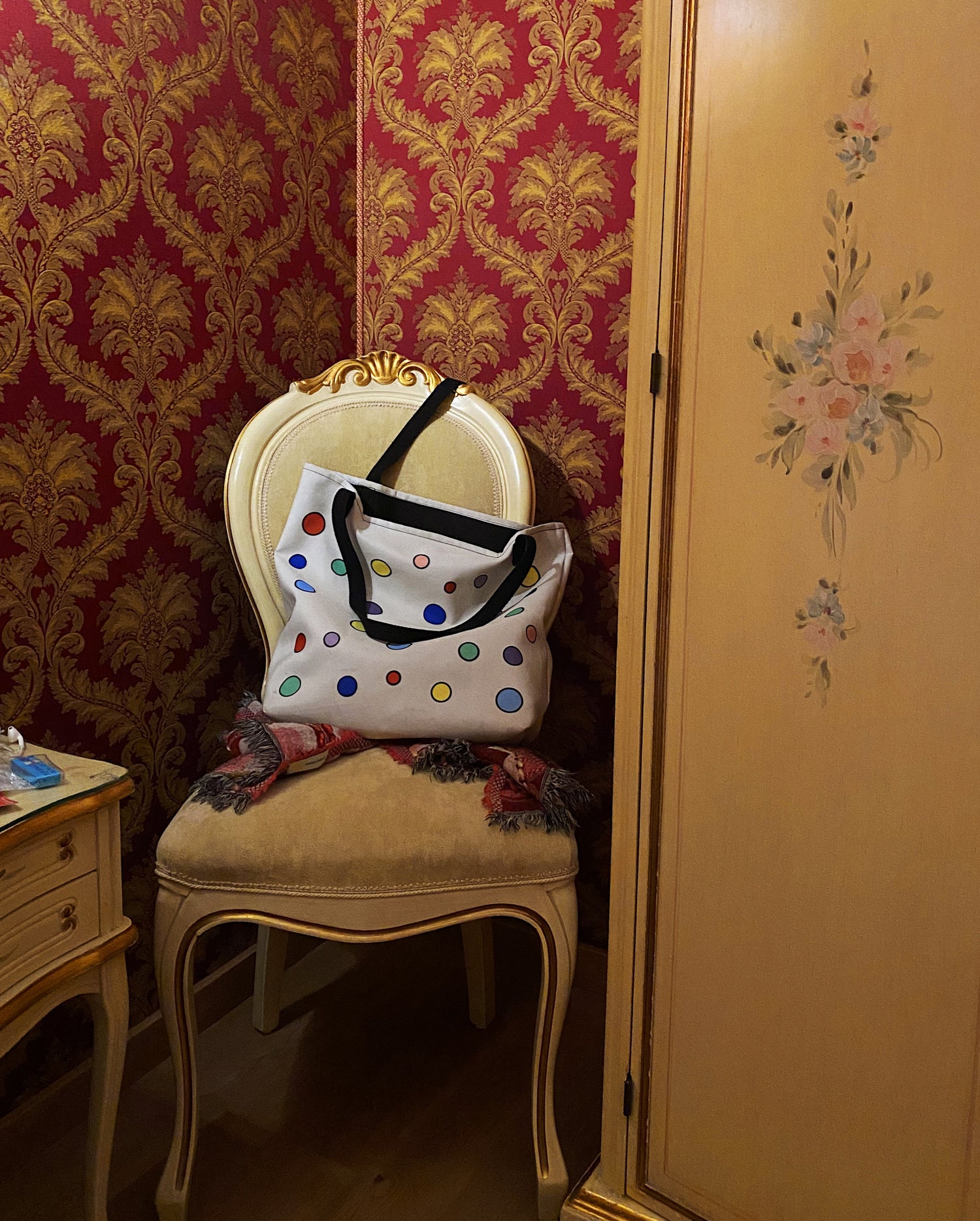 a colorfully polka-dotted cream colored tote bag sits slouched in an antique chair next to a hand-painted antique wardrobe in a richly wallpapered burgundy hotel room in Italy.