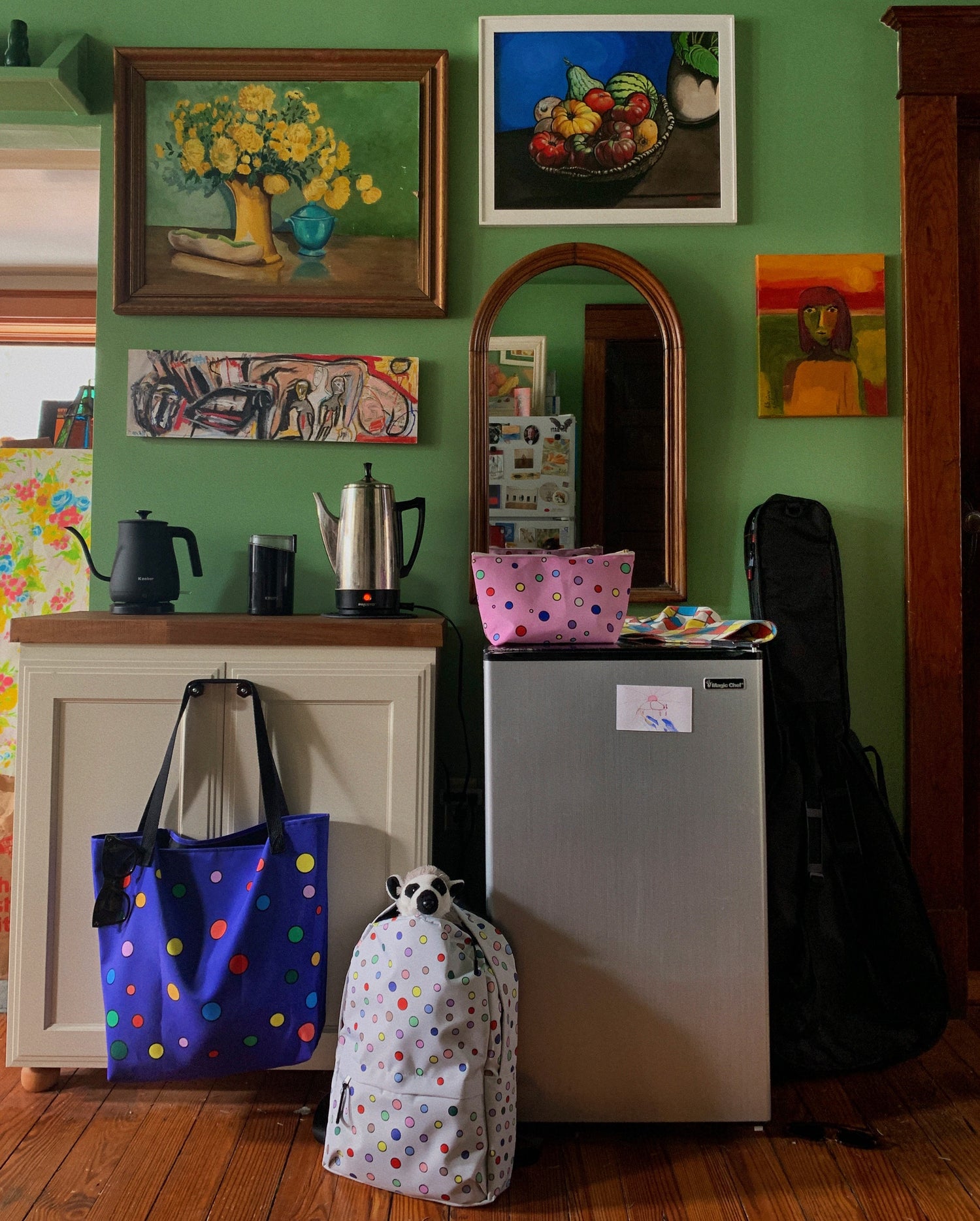 purple wizard bag, polka dotted sacred chemistry backpack, and atomic jubilee pink accessory pouch shown hanging and laying on a coffee bar with kettles and a mini fridge with a guitar case leaning on it, under an eclectic collection of still life and abstract paintings in a deep pistachio green kitchen with an antique arched mirror and classic wood doorway trim.