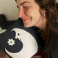 A smiling brunette holding and hugging the black and white lunar fool pillow against her chin, laying down in a bed.