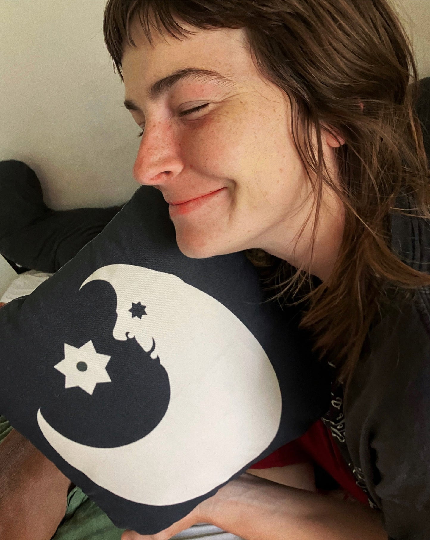A smiling brunette holding and hugging the black and white lunar fool pillow against her chin, laying down in a bed.