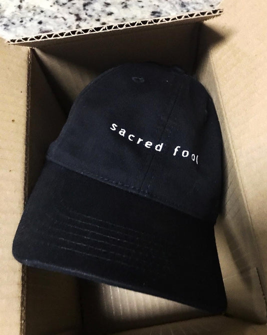 sacred foot black dad hat with white embroidered logo shown in open cardboard box. picture submitted by a happy customer!