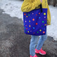 a brilliant purple tote bag with multicolor polka dots is held over the shoulder by a person in a yellow sweatshirt, denim pants, and flourescent pink nike sneakers in a driveway lined with 4 inches of visible snow and a row of evergreen trees.