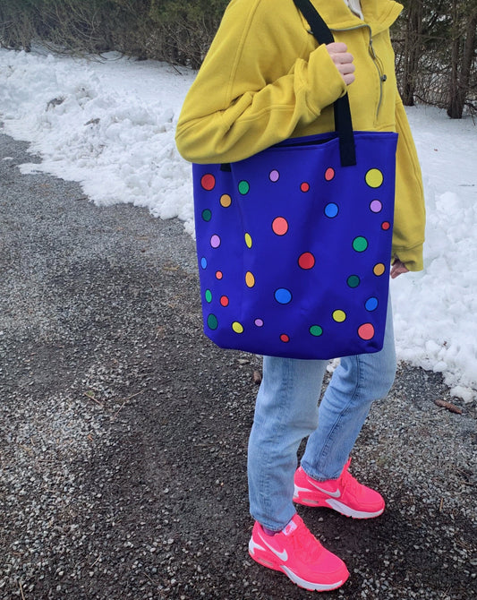 a brilliant purple tote bag with multicolor polka dots is held over the shoulder by a person in a yellow sweatshirt, denim pants, and flourescent pink nike sneakers in a driveway lined with 4 inches of visible snow and a row of evergreen trees.